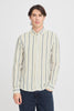 A person with long dark hair is casually dressed for the occasion in a Button Linen Cotton Shirt 20505463 by Casual Fridays, featuring white, beige, and blue vertical stripes. They stand with one hand in their dark jeans pocket against a plain white background.