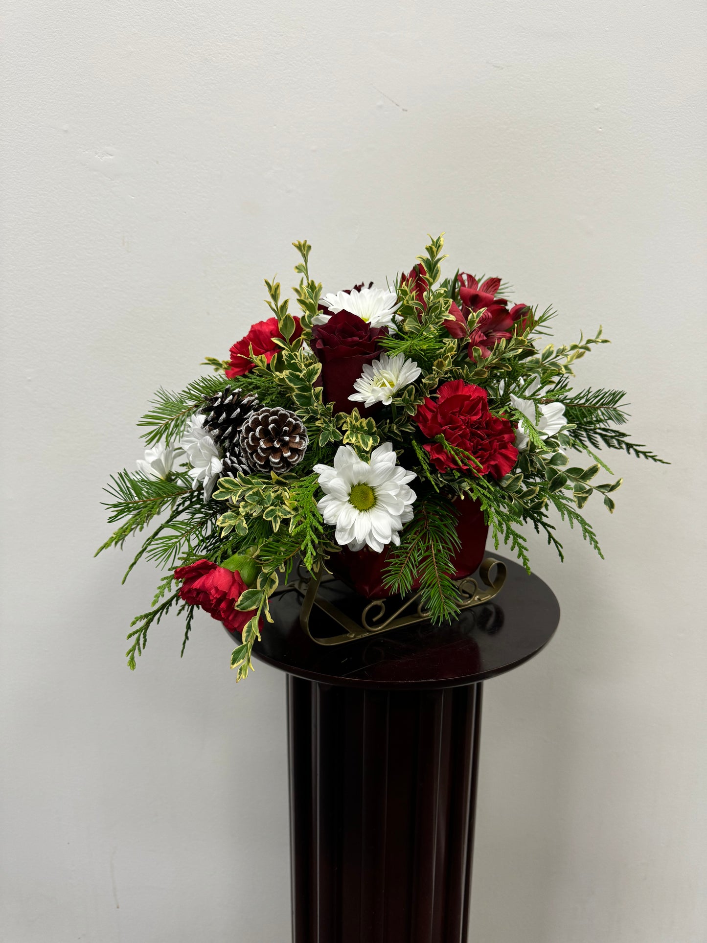 Introducing "Here Comes the Sleigh" by Posies Flowers & Fashion: a festive creation for the holiday season featuring a delightful combination of red and white blooms, vibrant green foliage, and charming rustic pinecones. This stunning arrangement is elegantly presented on a dark pedestal set against a clean light backdrop.