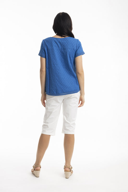 A woman faces away, wearing the ORIENTIQUE Essential Broderie Top (Style: 42097) in textured blue cotton with short sleeves, paired with white knee-length pants and beige heeled sandals against a plain white background.