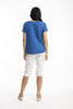A woman faces away, wearing the ORIENTIQUE Essential Broderie Top (Style: 42097) in textured blue cotton with short sleeves, paired with white knee-length pants and beige heeled sandals against a plain white background.