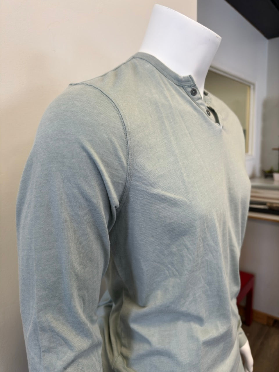 A mannequin dressed in the Silver Jeans Co. Shirt Long Sleeve - SAGE, featuring a classic 3-button design along the V-neckline. The sage green shirt, made from a comfortable cotton-polyester blend, is paired with black pants. The background is plain and white.