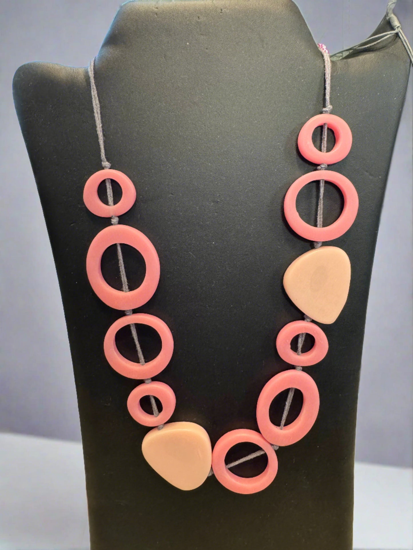 The Necklace Adjustable - PINK by Suzie Blue, displayed on a black stand, features an array of various sized and shaped beads threaded onto a dark string. The circular, oval, and teardrop beads come together to create a striking piece of statement jewelry that is both stylish and versatile.