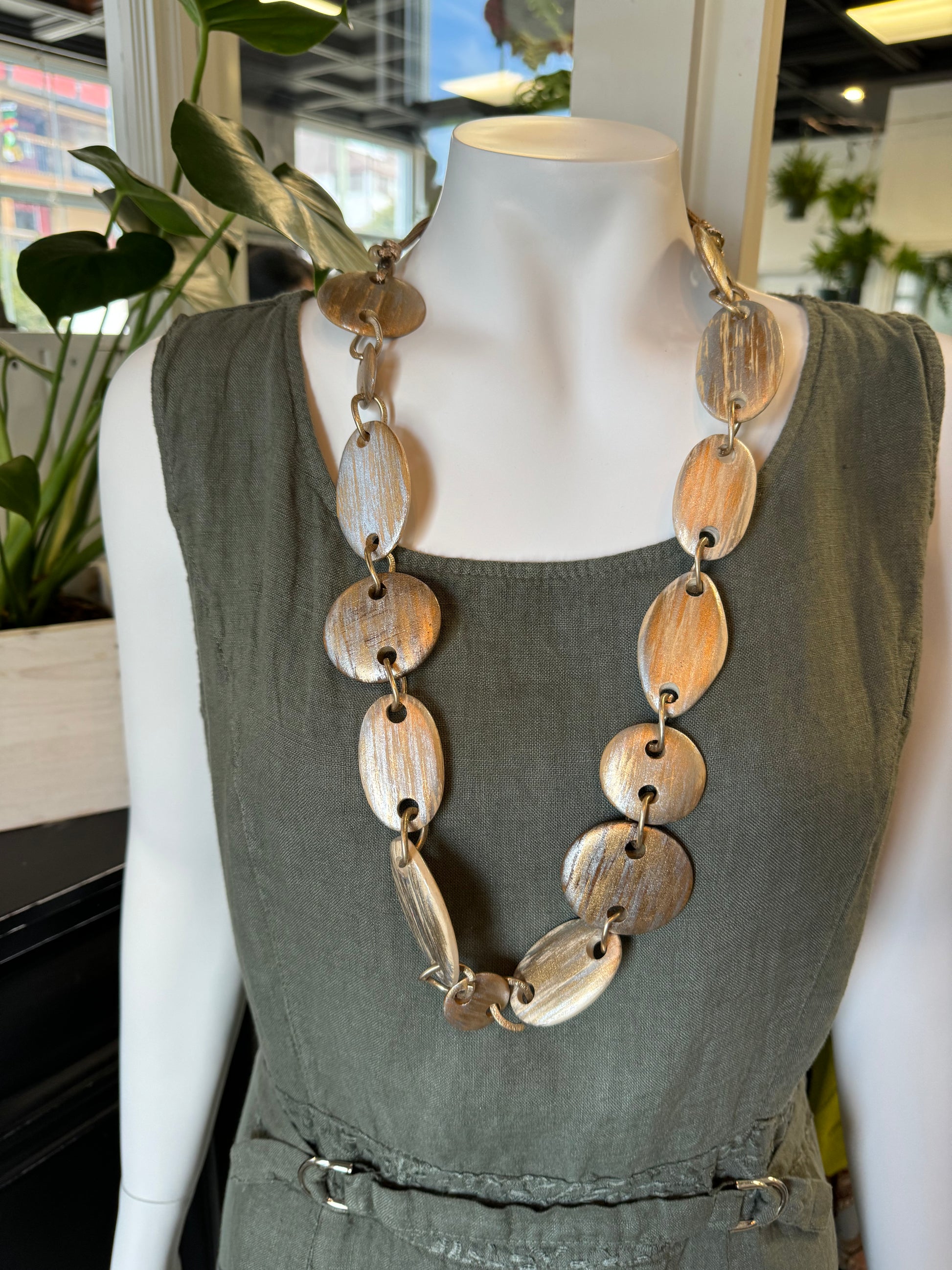 A mannequin dressed in a sleeveless, sage green dress is adorned with an elegant Silver and Gold Coloured Necklace from Posies Flowers & Fashion Inc. The background features potted plants and a modern indoor space.