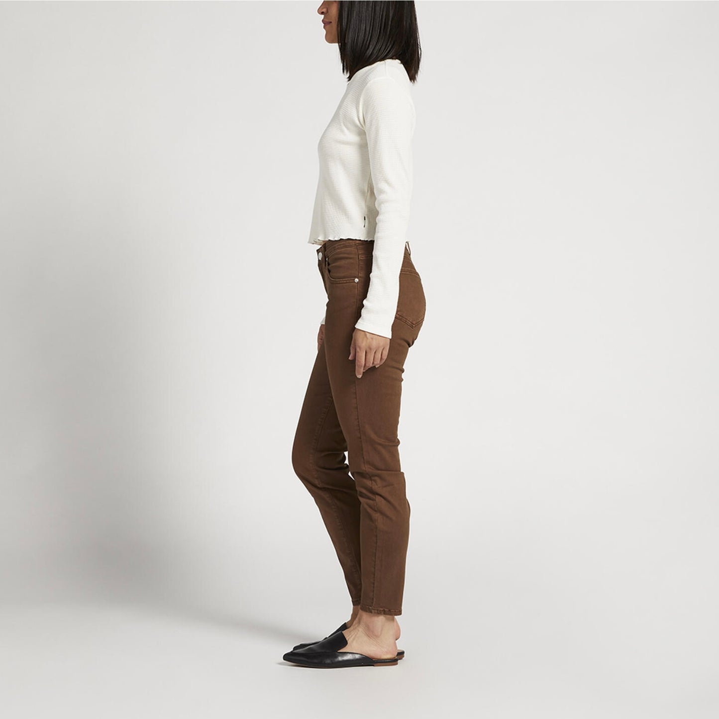 A person wearing a long-sleeved white shirt, Cecilia Mid Rise Skinny Pants - JAG in brown, and black slip-on shoes stands against a plain white background. They are slightly turned to the right and have one arm resting casually by their side.