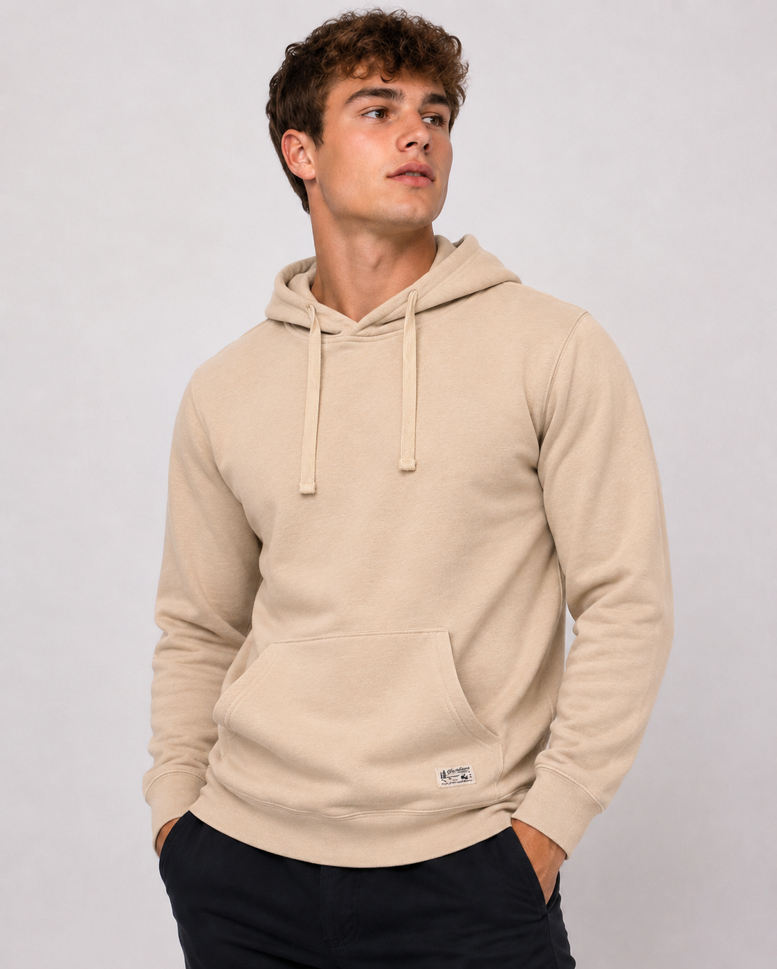 A young man stands against a plain light background wearing a light beige Northbound Supply Co Hoodie with a front pocket and drawstrings, looking slightly to the side with his hands in his pockets.