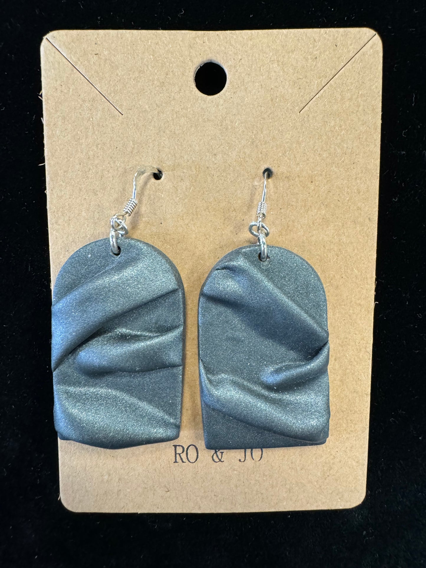 A pair of metallic gray handmade earrings displayed on a brown kraft paper card against a dark background. The earrings feature a wavy, textured design and hang from silver hooks. The card has a hole at the top for hanging and text that reads "RO&JO Earrings 438" at the bottom.