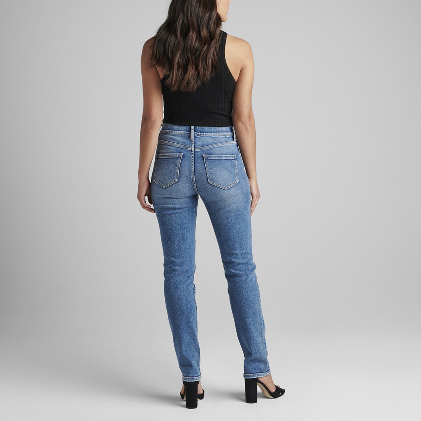 A person stands against a plain gray background, wearing a black sleeveless top and the Valentina High Rise Straight Leg Pull-On Jeans from JAG. They have long, wavy dark brown hair, and are wearing black open-toe heels with their arms relaxed by their sides.