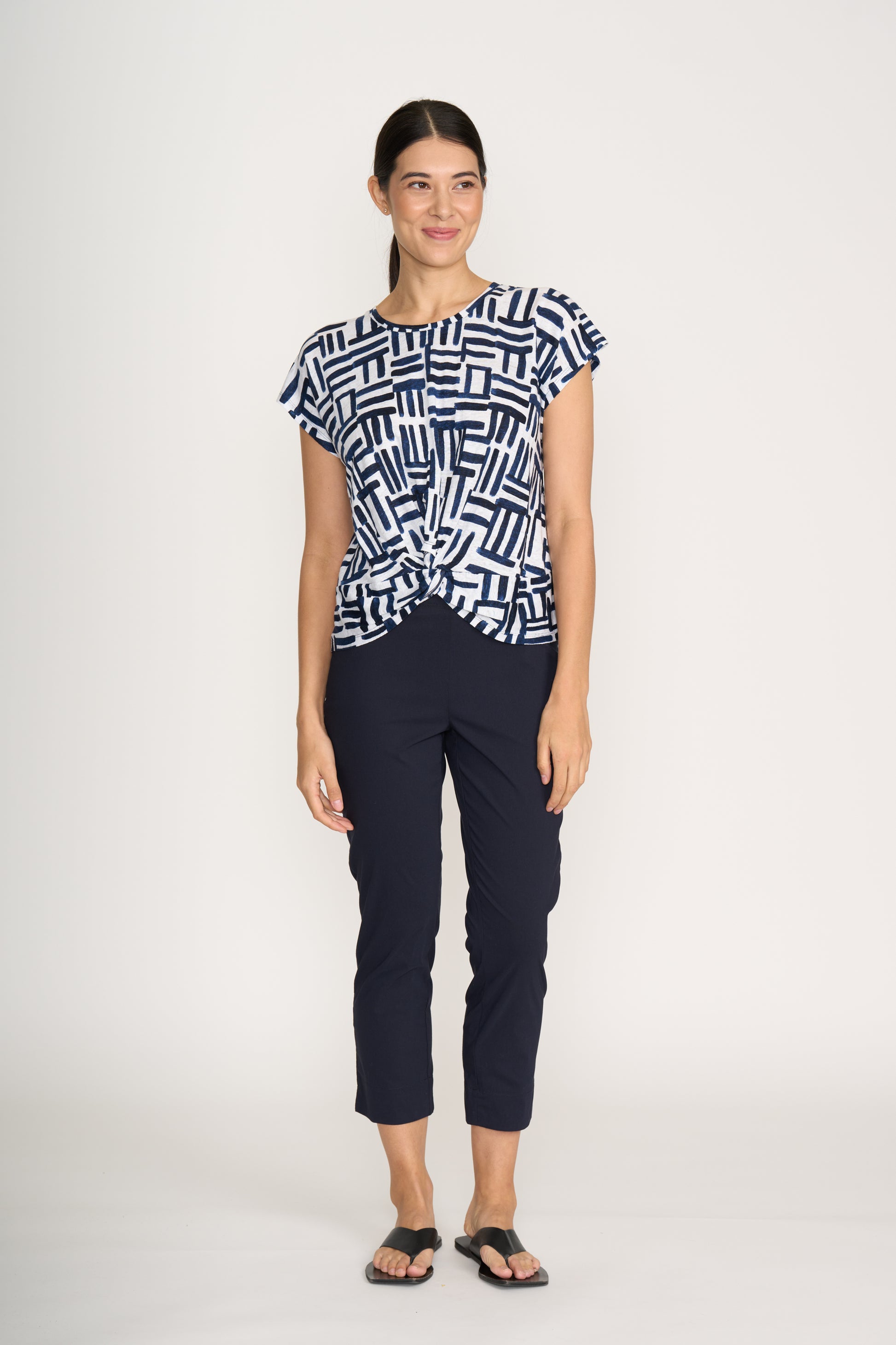 A woman stands against a plain background, smiling with one hand by her side, wearing the FOIL Do The Twist Top FO8258 Block & Roll paired with navy cropped pants and black flip-flops.