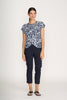 A woman stands against a plain background, smiling with one hand by her side, wearing the FOIL Do The Twist Top FO8258 Block & Roll paired with navy cropped pants and black flip-flops.
