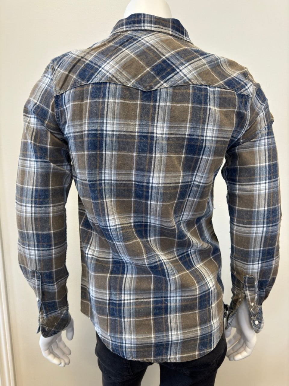 A mannequin wears the Shirt Long Sleeve - OLIVE by Silver Jeans Co., showcasing a plaid design in shades of brown, blue, and white. Crafted from 100% cotton, this button-up shirt features two front chest pockets with buttoned flaps. The mannequin's hand is partially visible on the right side as it stands against a plain, light-colored background.