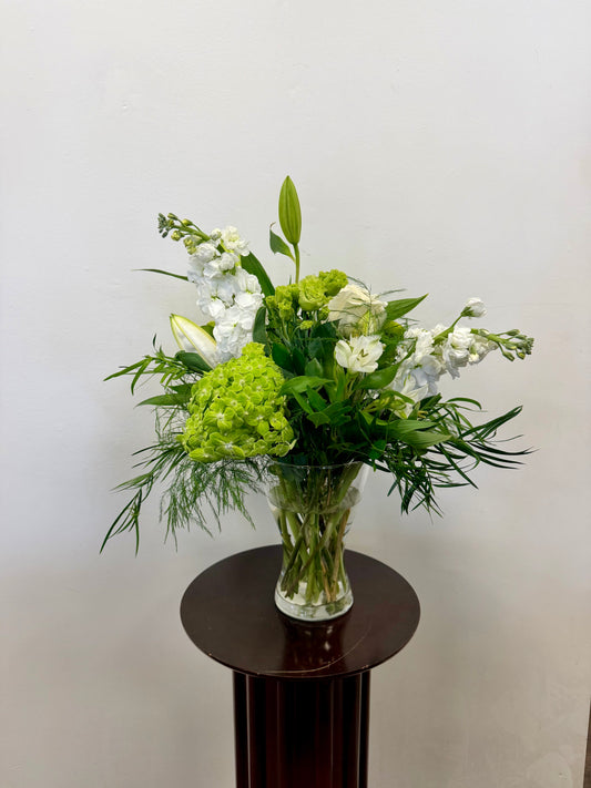 The Whites and Greens Vase by Posies Flowers & Fashion, featuring assorted green and white flowers with leafy greenery, is displayed on a small round dark wooden table against a plain white wall.