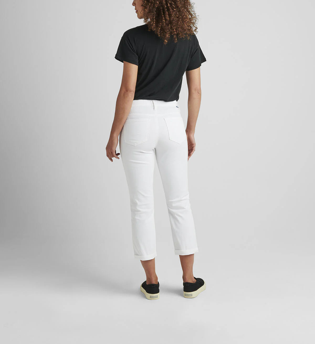 A person with curly hair, wearing a black t-shirt and the Cecilia Mid Rise Capri White from JAG, paired with black sneakers with white soles, is standing and facing away from the camera. The background is plain and gray.