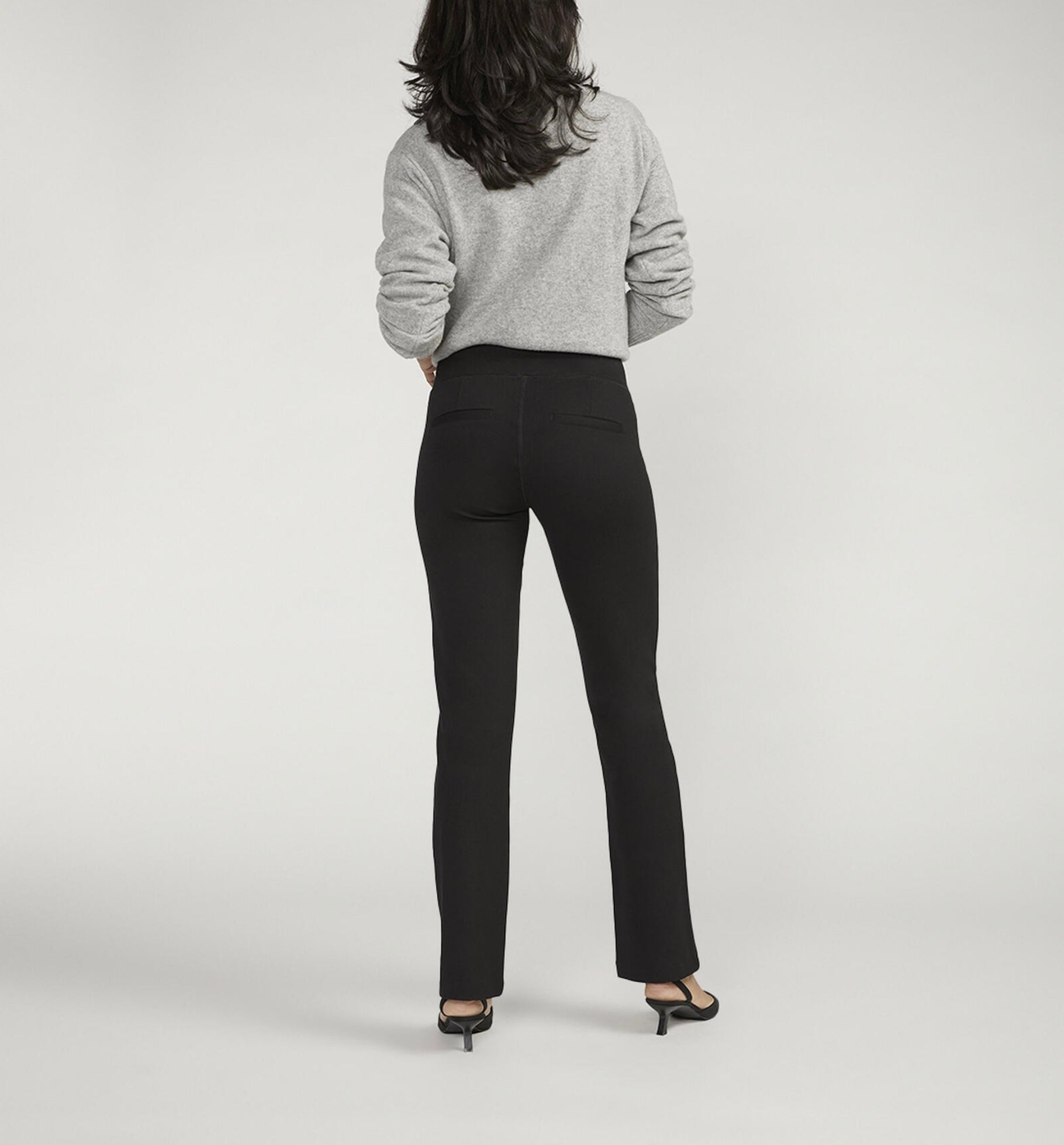 A person is posing against a plain background, wearing a light gray long-sleeve sweater paired with JAG's Mid Rise Bootcut Pull-On Pants. They have one hand on their hip and are sporting black pointed-toe heeled shoes. Their dark hair flows loose and wavy.