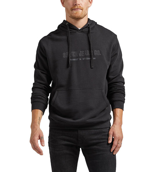 A person is wearing the Silver Jeans Co. Logo Hoodie in black, featuring "Silver Jeans Co." embroidered on the front along with geographic coordinates. The hoodie includes a front pocket and drawstrings. They are also sporting black jeans and have light facial hair.