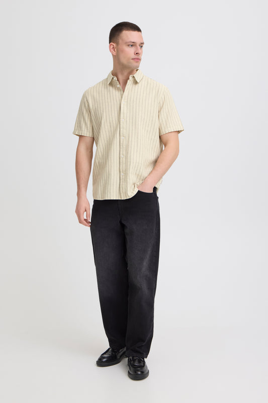 A man stands against a plain white background wearing the BLEND BHAKI SHIRT 20720666-900007166, paired with loose black jeans and black shoes. He looks to his left with one hand in his pocket.