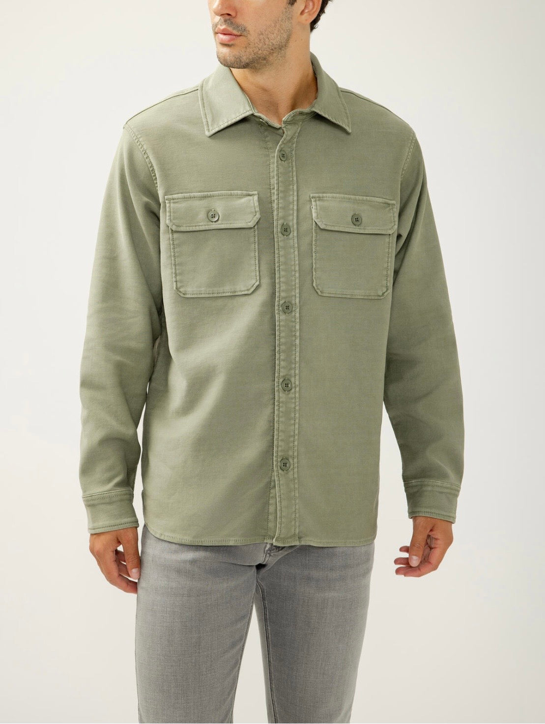 A man stands against a plain background wearing the Silver Jeans Co. Knit Flex Button-Up Shirt MJ0035KTS674, a relaxed fit light olive green shirt with two chest pockets, styled with light gray jeans and his head slightly turned to the side.