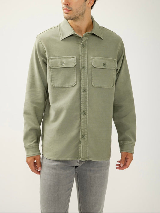 A man stands against a plain background wearing the Silver Jeans Co. Knit Flex Button-Up Shirt MJ0035KTS674, a relaxed fit light olive green shirt with two chest pockets, styled with light gray jeans and his head slightly turned to the side.