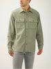 A man stands against a plain background wearing the Silver Jeans Co. Knit Flex Button-Up Shirt MJ0035KTS674, a relaxed fit light olive green shirt with two chest pockets, styled with light gray jeans and his head slightly turned to the side.