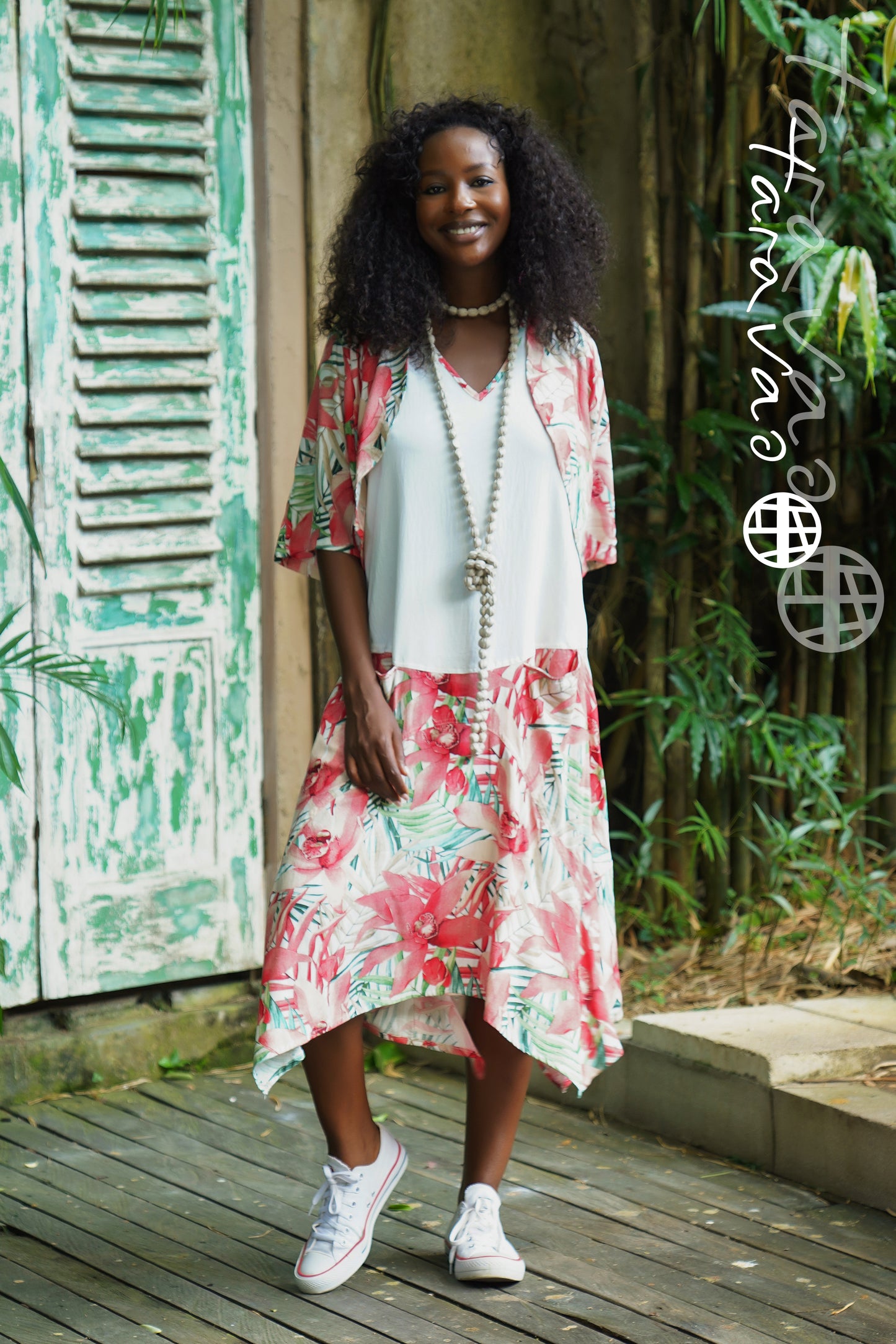 A smiling woman with curly hair wears the TaraVao Dress Megan 8153, a rayon dress with red and green floral print, paired with a beaded necklace and white sneakers. She stands by distressed green shutters and lush plants outdoors.