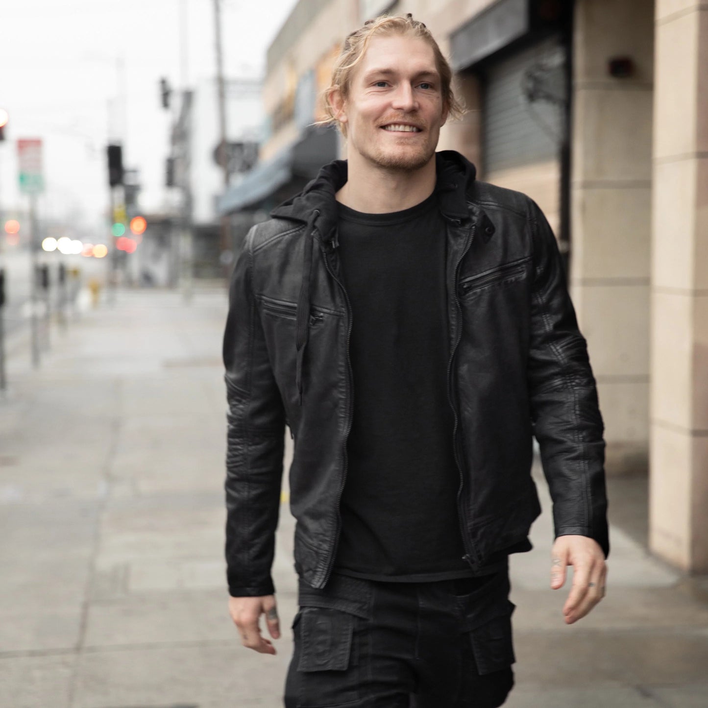 A person with light hair is walking down a city street, wearing the Kayto RF Leather Jacket in black from MAURITIUS LEATHER over a black shirt and black pants. The background is slightly blurred, showing buildings, a sidewalk, and streetlights. The person is smiling and appears to be enjoying the walk.