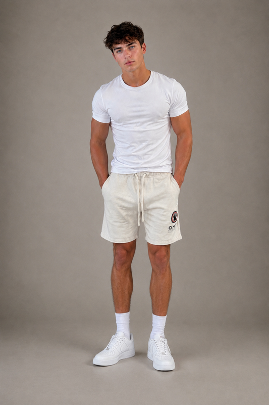A man stands against a plain gray background wearing a fitted white t-shirt and Once We Were Warriors Yabu Shorts in WHITE SAND, paired with white crew socks and sneakers. His hands are in his pockets as he faces forward.