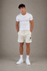A man stands against a plain gray background wearing a fitted white t-shirt and Once We Were Warriors Yabu Shorts in WHITE SAND, paired with white crew socks and sneakers. His hands are in his pockets as he faces forward.