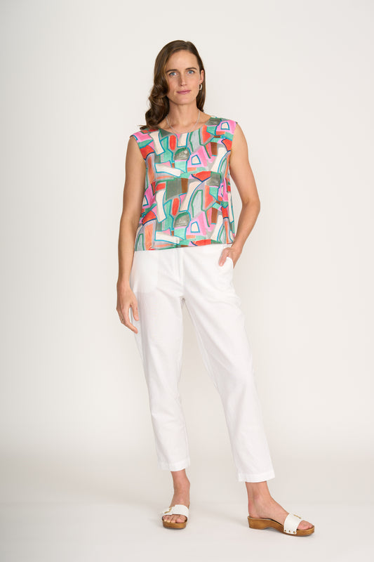 A woman stands against a plain white background wearing the FOIL Its Only Natural Top FO6917 Colour Fusion, paired with white pants and sandals. She has one hand in her pocket and a neutral expression.
