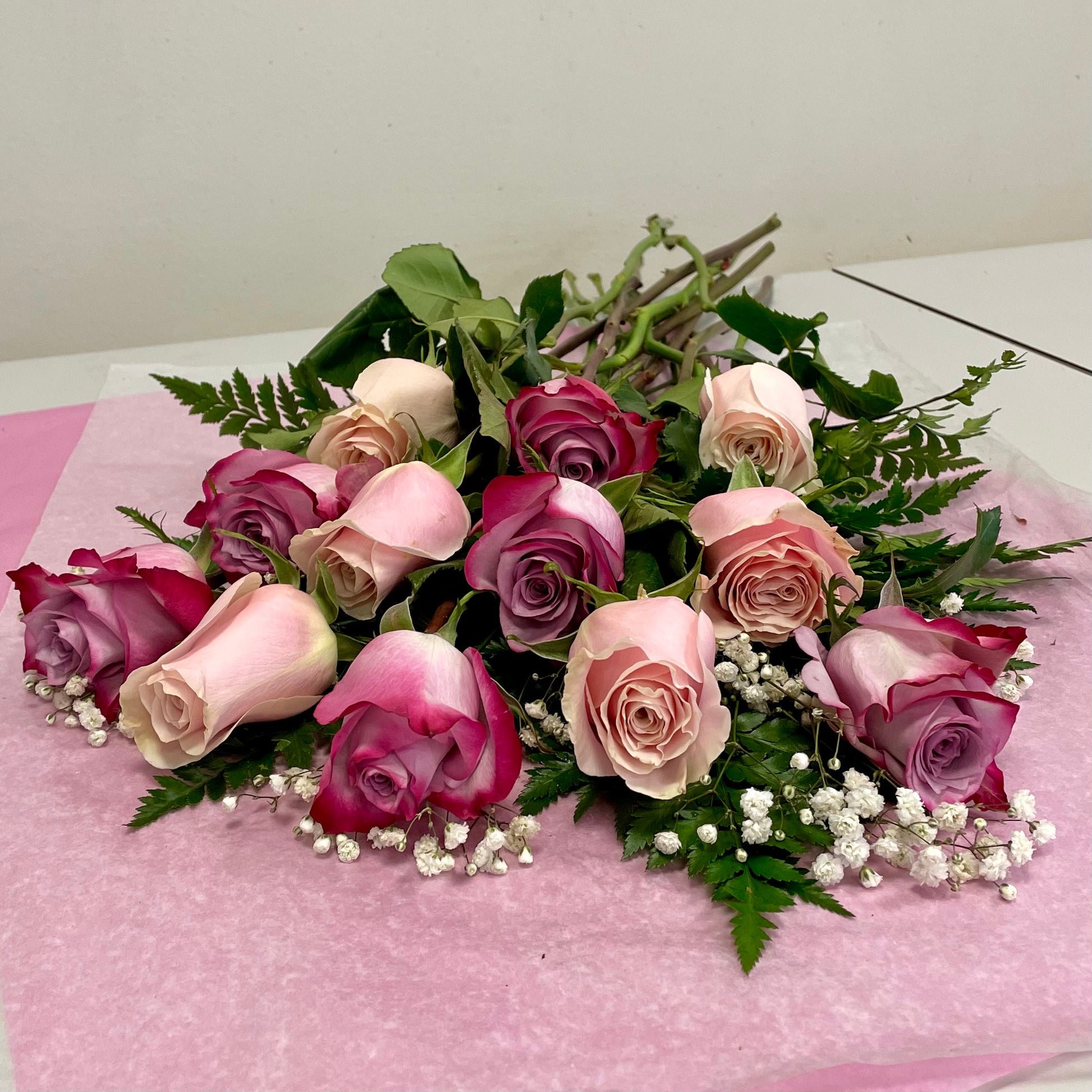 The 1 Dozen Mixed Pink Roses Freshcut from Posies Flowers & Fashion features pink and purple roses with baby's breath and green fern leaves, elegantly arranged on pink tissue paper.