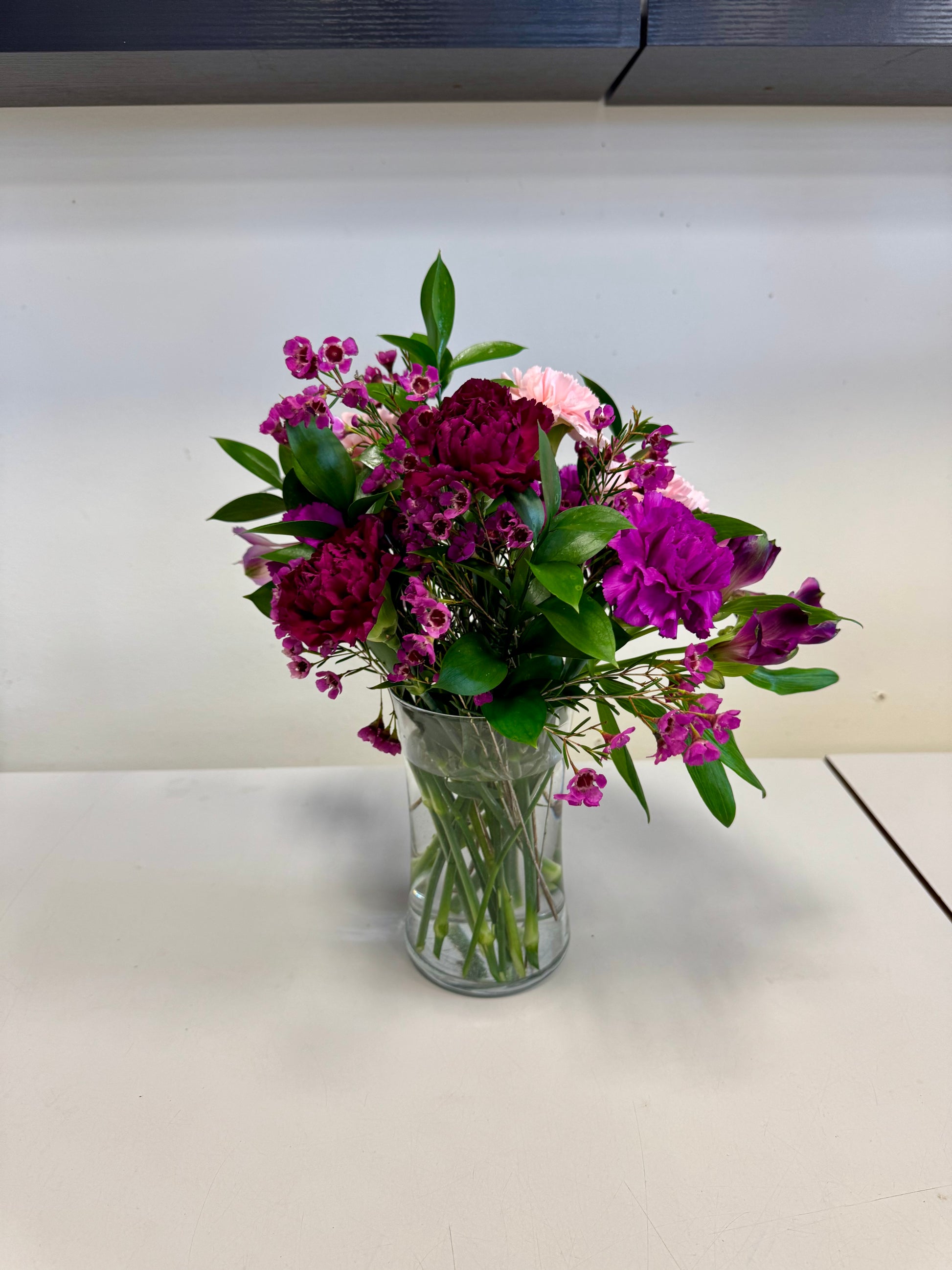 The "Based in Pink" arrangement by Posies Flowers & Fashion features magenta, purple, and pink blooms with green leaves in a glass vase on a white surface; substitutions may occur due to seasonal availability.