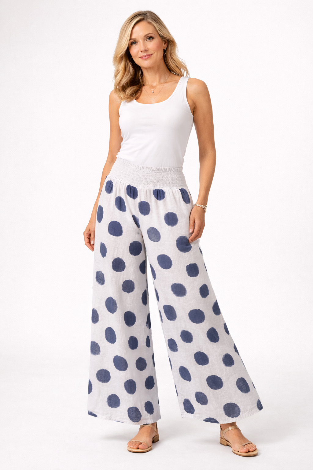 A woman with long blonde hair stands confidently against a white background, wearing Bella Amore Linen Pant 30201 White and a sleeveless top, paired with sandals. Made in Italy, her look is relaxed and stylish.