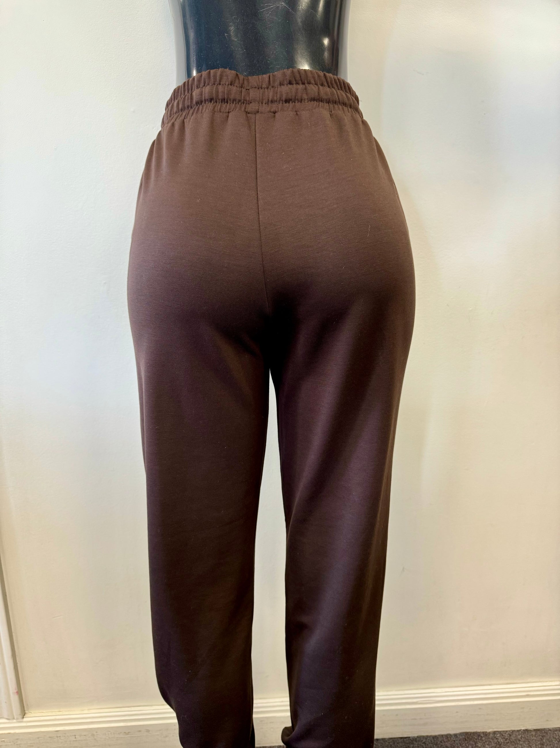 The image shows a person wearing SoyaConcept Banu 117 Relaxed Drawstring Pant in a brown jogger-style with a customizable fit, featuring a drawstring waist and elastic cuffs at the ankles, perfect for any casual occasion. They're standing against a plain light-colored wall.