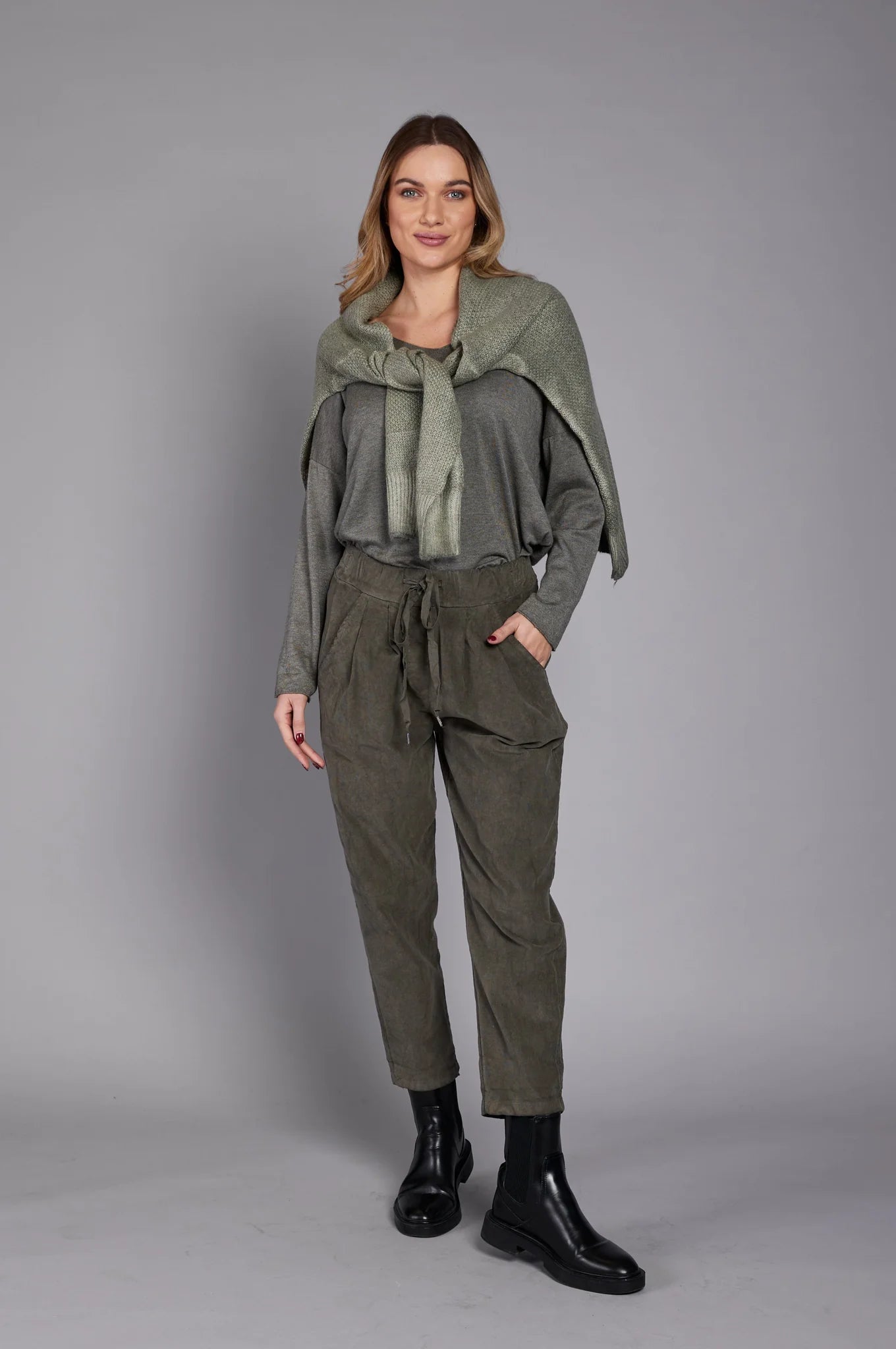 A person wearing a gray long-sleeve top, the stylish EL27901 pants by ELISSIA in ankle-high olive green with a drawstring, and black ankle boots stands with their hands in their pockets against a plain grey background. A scarf is draped over their shoulder. Only the lower half of the person is visible.