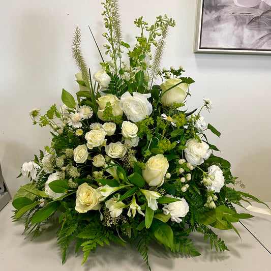 The "Whispering Elegance" by Posies Flowers & Fashion is a grand funeral tribute showcasing white roses, lilies, and assorted green foliage. These delicate flowers are meticulously arranged in an elegant, symmetrical design that results in a lush and full display. The backdrop features a white wall with a partially visible framed picture.