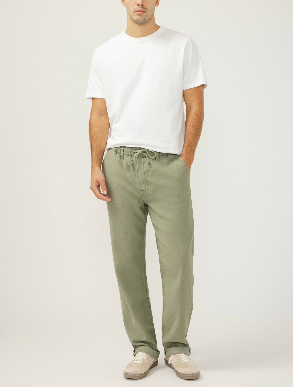 A person stands against a plain background wearing a white short-sleeve t-shirt, Silver Jeans Co. Relaxed Fit Trouser Leg Knit Flex Jeans (M43418KTS674), and light-colored sneakers, hands in their pockets.