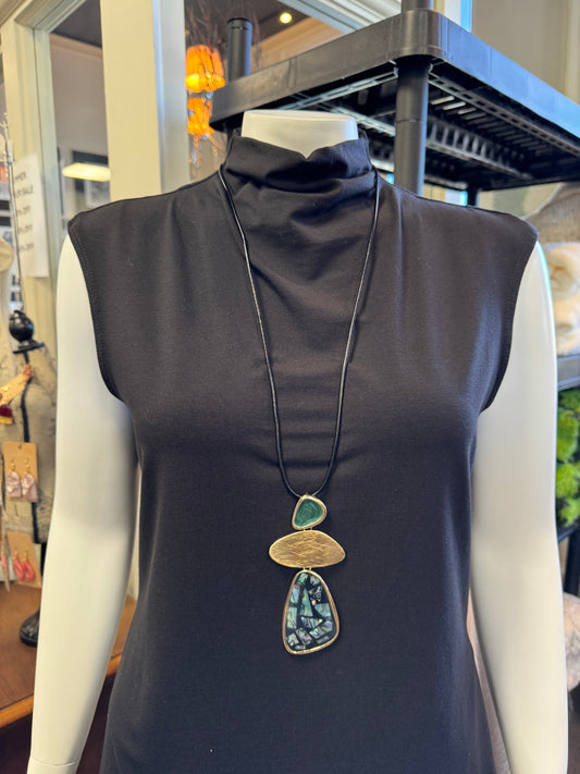 A mannequin features a sleeveless black dress highlighted by a striking accessory: Necklace 9 from Elise Accessories. The large pendant in green and blue shades gracefully hangs on a black cord, making it a fashion-forward statement piece. In the background, the shop interior displays an assortment of trendy jewelry and other stylish items.