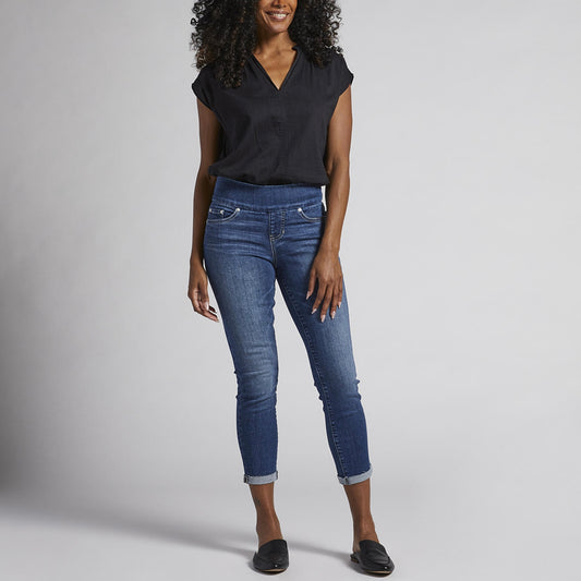 A person with curly hair is wearing a black short-sleeve blouse, Amelia Mid Rise Slim Ankle Pull-On Jeans by JAG, and black loafers. They are standing against a plain light grey background, smiling, with their hands relaxed by their sides.