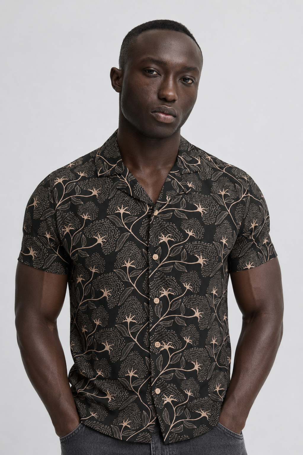 A man stands against a plain background wearing the BLEND ARIAN SHIRT 20720641-099996162, a casual short sleeve men's shirt with a dark floral pattern in soft viscose. His hands are in his pockets and he has a neutral expression.