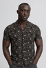 A man stands against a plain background wearing the BLEND ARIAN SHIRT 20720641-099996162, a casual short sleeve men's shirt with a dark floral pattern in soft viscose. His hands are in his pockets and he has a neutral expression.