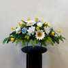 The 1 - Tombstone Saddle (60 cm) by Posies Flowers & Fashion Inc. features white daisies, yellow tulips, light blue roses, and green ferns on a round black stand against a plain light wall.