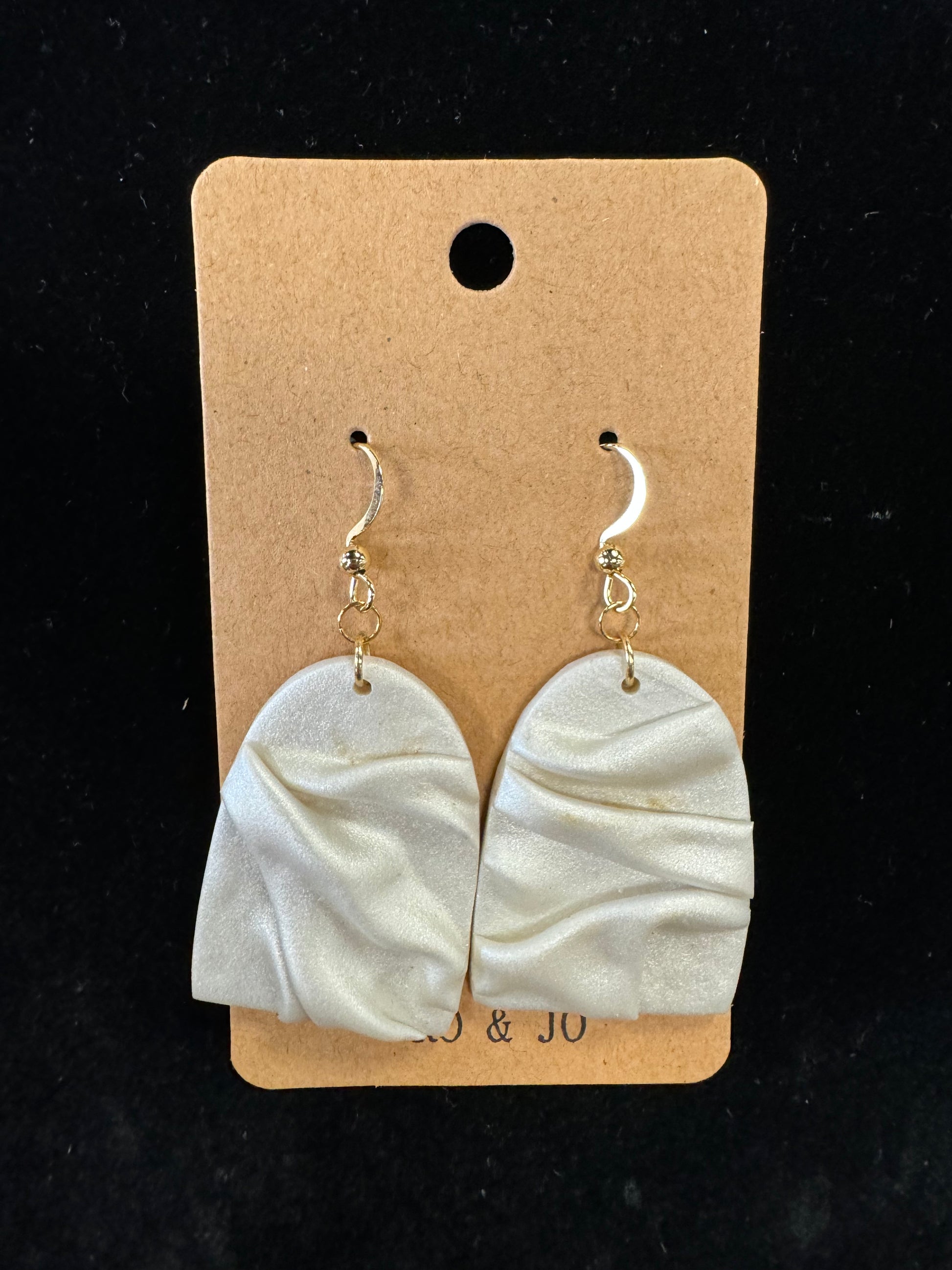 The Ro&Jo Earrings 442 by RO&JO are displayed on a tan card. These artisan crafted, handmade earrings feature white, draped polymer clay pendants with gold hook clasps. The black background provides a striking contrast to both the earrings and the card.