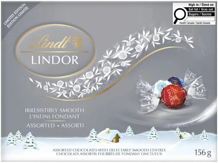 Lindt Lindor Assorted Chocolate Truffles Box, 156g, comes in winter-themed packaging with snow, trees, and cabins. Three foil-wrapped Swiss chocolates are shown on the right. This Limited Edition box is perfect for gifting.