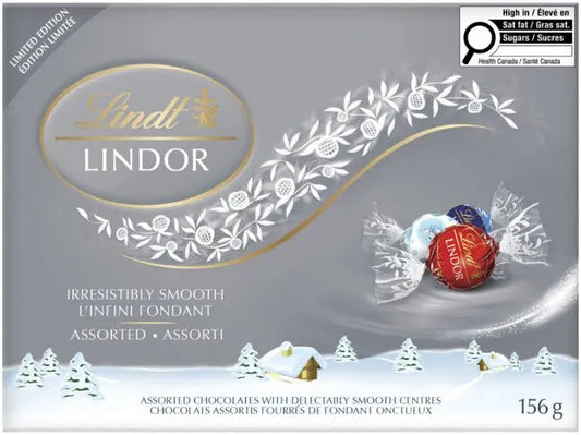 Lindt Lindor Assorted Chocolate Truffles Box, 156g, comes in winter-themed packaging with snow, trees, and cabins. Three foil-wrapped Swiss chocolates are shown on the right. This Limited Edition box is perfect for gifting.