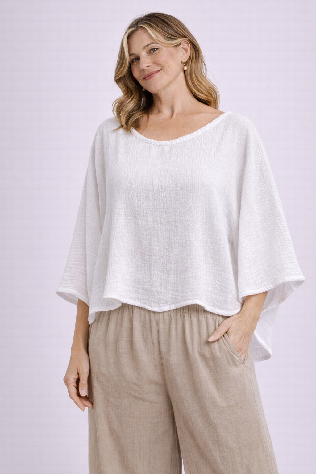 A woman with wavy hair wears the Oh My Gauze Vanna Snow Blouse T295 and beige wide-legged pants, standing and smiling with one hand in her pocket against a light, plain background.