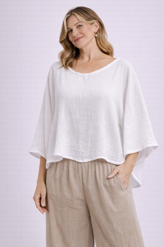 A woman with wavy hair wears the Oh My Gauze Vanna Snow Blouse T295 and beige wide-legged pants, standing and smiling with one hand in her pocket against a light, plain background.