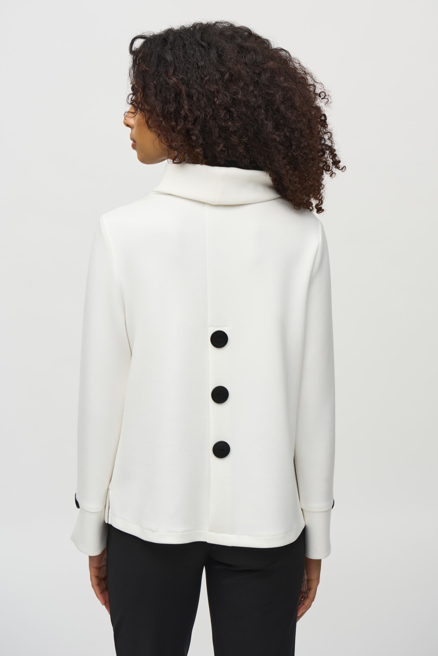 A woman with curly hair wears a loose-fitting, long-sleeved white Scuba Jersey Boxy Top 244129 by Joseph Ribkoff. The top, featuring button details on the sleeves, lends a boxy silhouette. She stands in front of a plain background, looking slightly downward with a neutral expression.