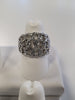 The Jenna-39 Adjustable Ring by Elise Accessories, a sophisticated silver-colored ring adorned with multiple rows of small, sparkling stones, is displayed on a white ring holder. The intricate design features interlocking loops encrusted with the stones, creating a wide and elegant band. The background is blurred.
