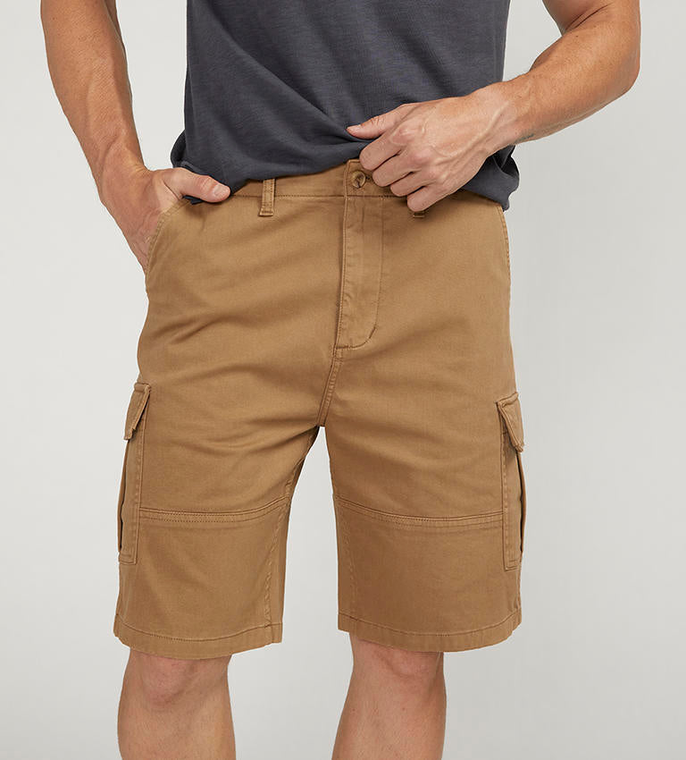 A man stands against a plain background wearing a dark gray t-shirt with a chest pocket, relaxed fit CARGO SHORT DESERT styled by Silver Jeans Co. (product code: M53151CBT639), and black sneakers with white soles. He appears to be adjusting his shirt with his right hand.