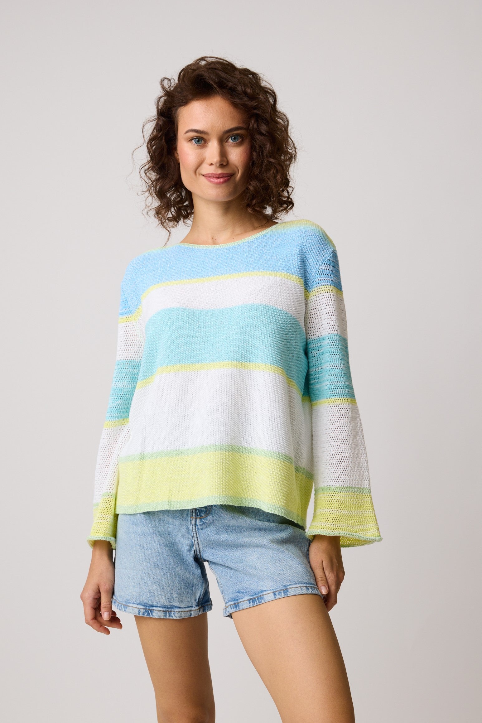 A person with curly hair smiles, donning a vibrant Parkhurst Naples Stripes Island Sky 80117 Cotton Country sweater in blue, green, yellow, and white. Paired with denim shorts, they stand against a plain light grey background.