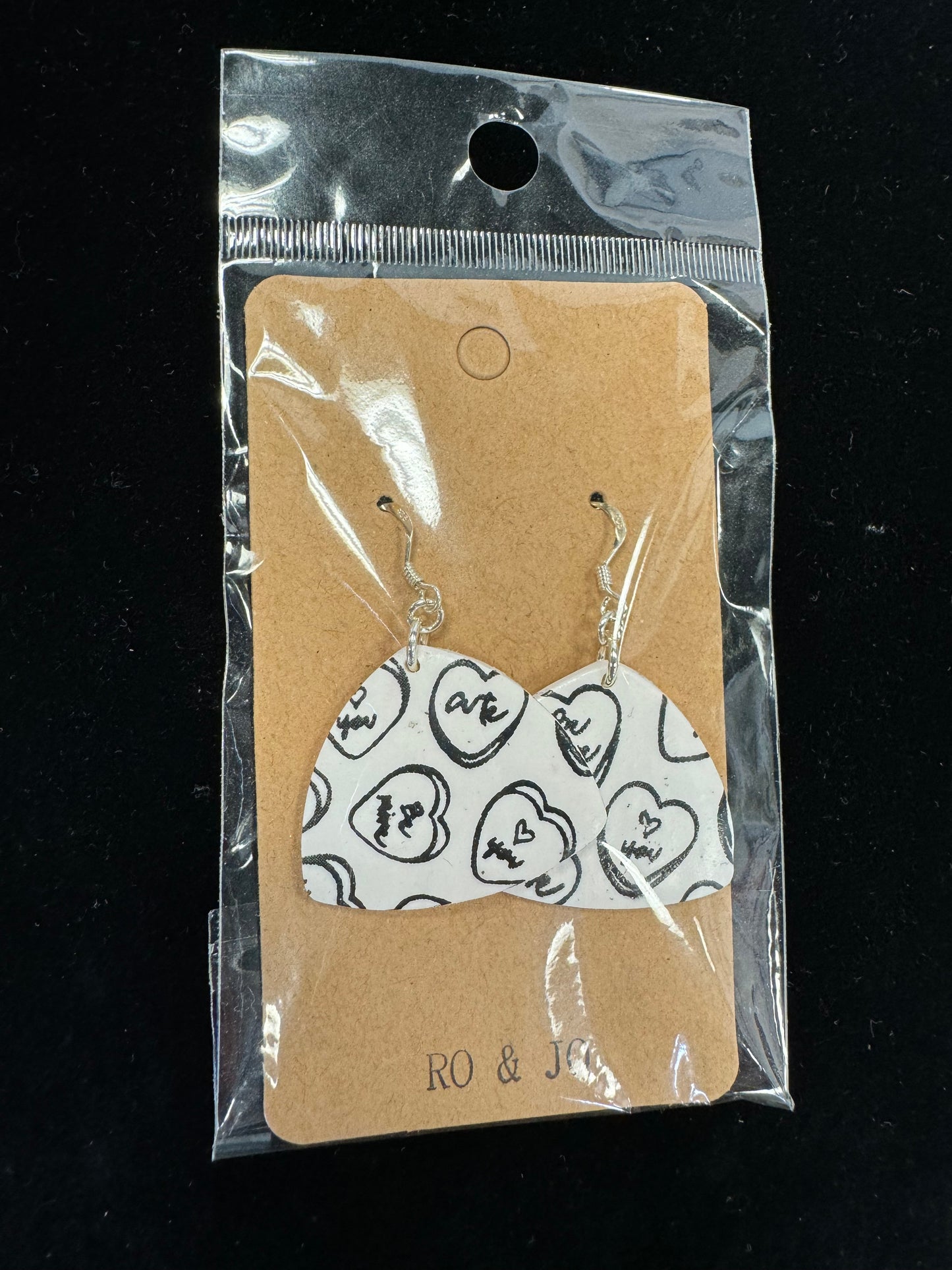 A pair of Ro&Jo Earrings 434, heart-shaped and handmade with a black and white design, are attached to a brown backing card that reads "RO&JO." Crafted from polymer clay, the earrings are packaged in a clear plastic sleeve. The background is black.