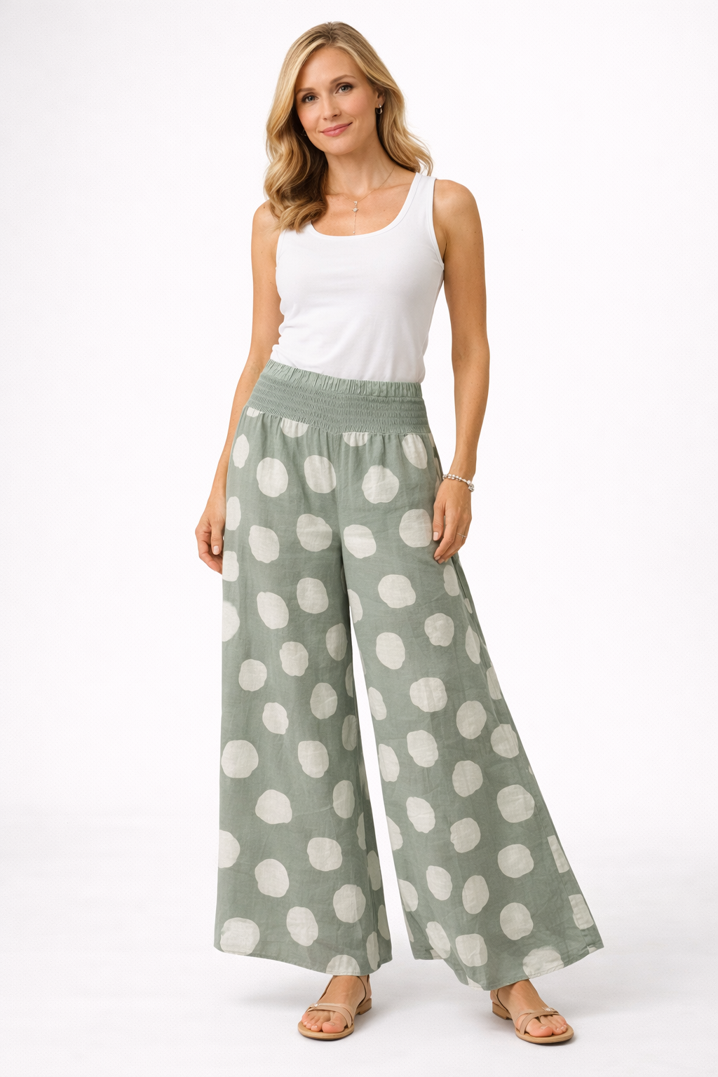 A woman with long, wavy blonde hair stands against a white background wearing the Bella Amore Linen Pant 30201 Sage—wide-leg, sage green linen trousers with large white polka dots—paired with a white sleeveless top and sandals. Made in Italy.